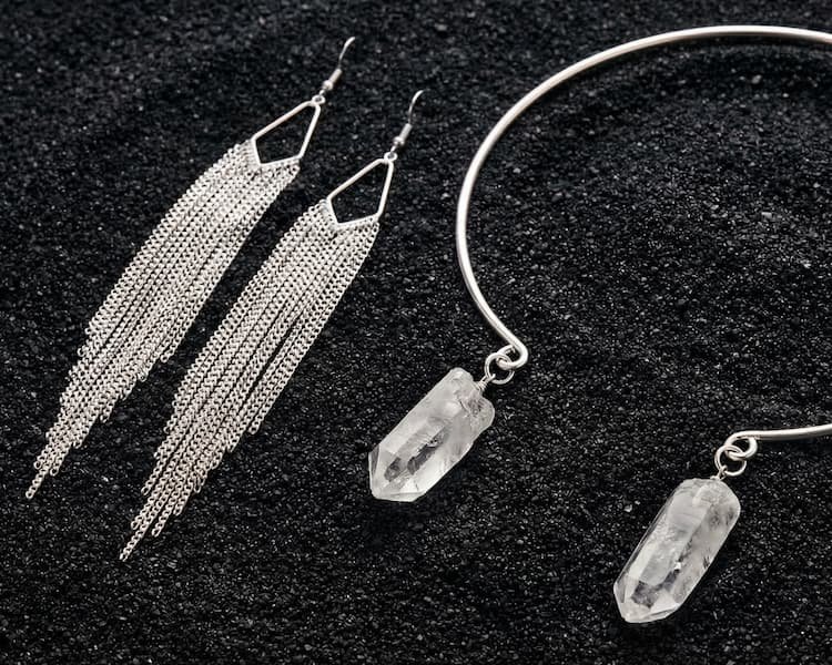 Minimalist metal jewelry composition featuring a silver neck torque with raw crystals and long industrial-style fringe earrings, representing a fusion of steel, silver, and bronze textures.